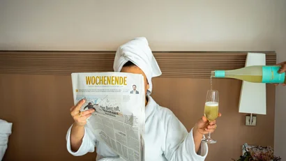 Person im Bademantel liest Zeitung, während Sekt in Glas eingeschenkt wird