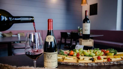 Rotwein wird in ein Glas gegossen neben Weinflaschen und einer Platte mit Käse und Obst