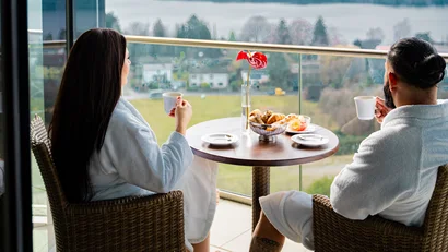 Paar in Bademänteln trinkt Kaffee auf Terrasse mit Blick auf See und Landschaft